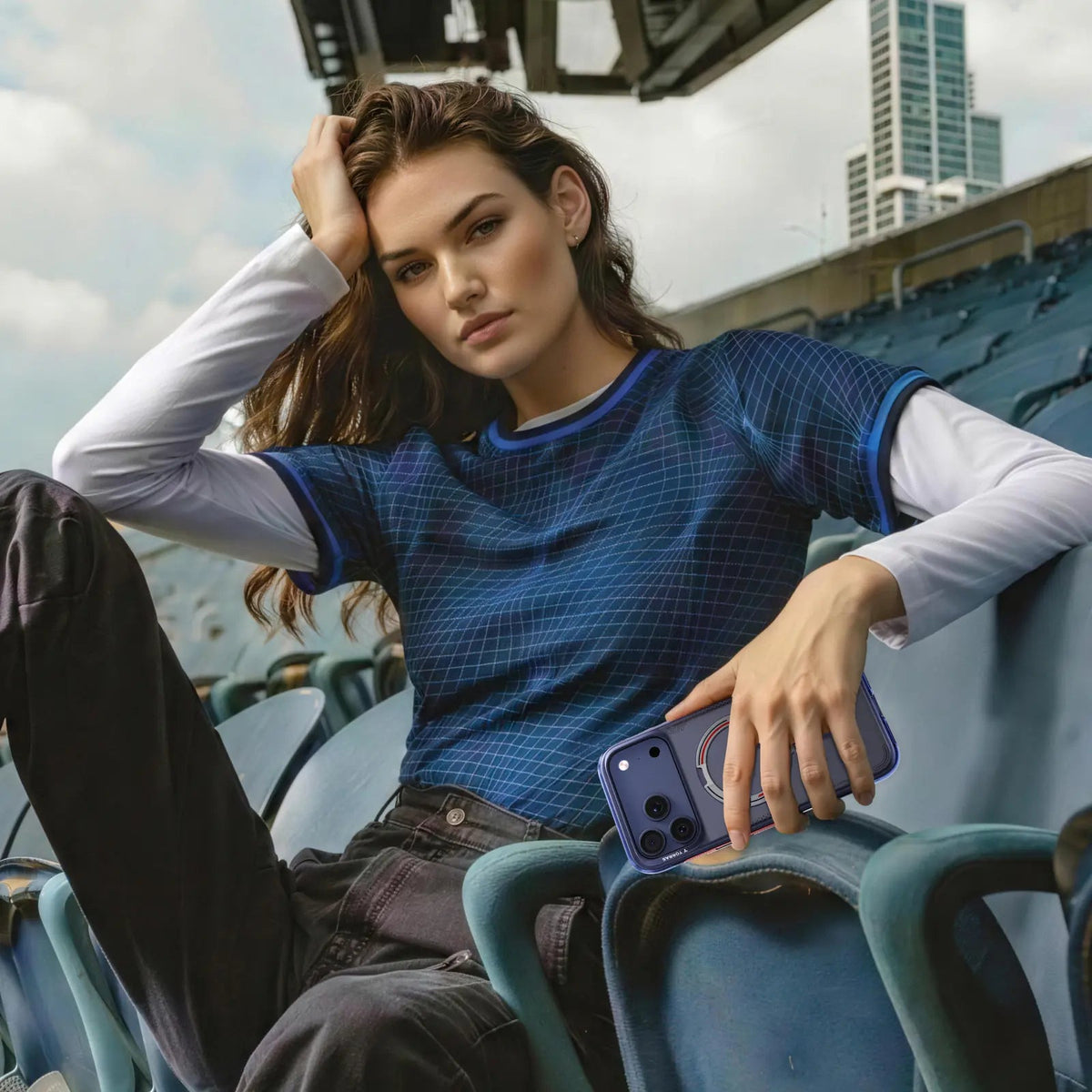 Woman in a blue football shirt in stadium seats, holding an iPhone 18 Pro Max in the blue Football case